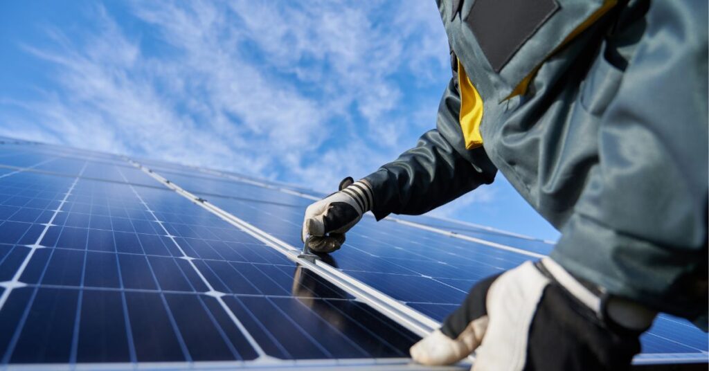 A person wearing a green jacket and gloves is installing a solar panel under clear blue skies.